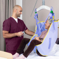 A woman is being lifted from her bed with assistance from a caregiver, using the Maxi Sky 2 lift thumbnail