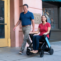 A Caucasian man and woman are out for a stroll on a city street. The woman uses a WHILL folding power chair. They are very happy. thumbnail