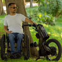 A man using a manual wheelchair sits in a verdant park. His Batec Scrambler electric handbike is detached, positioned beside him. thumbnail