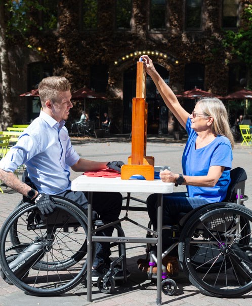 Two people using SmartDrives playing a board game at an outdoor table in the sunshine.
