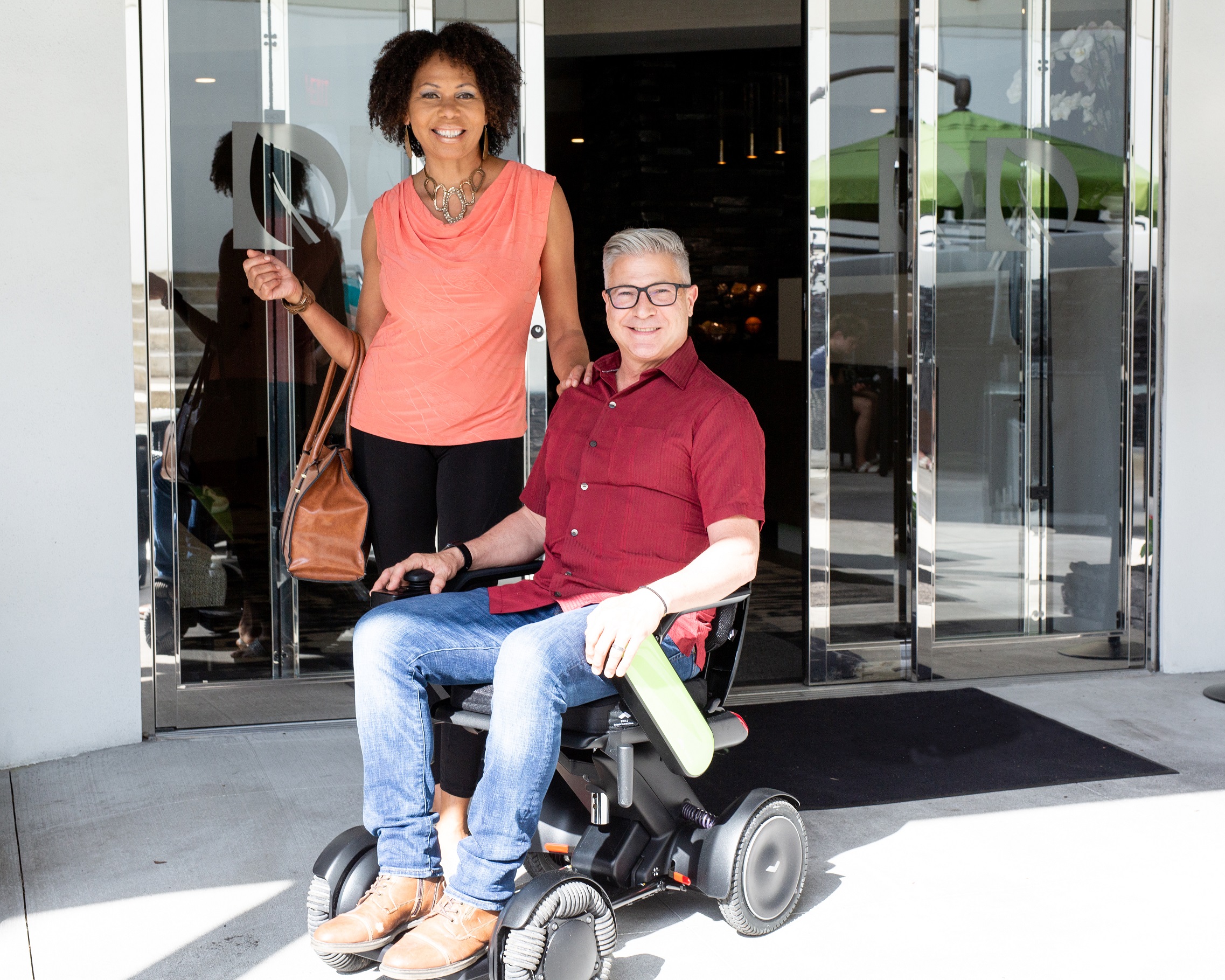 WHILL C2 couple in front of sliding doors A man sits in a white WHILL C2 power chair with a woman standing beside him. They are both smiling at the camera.