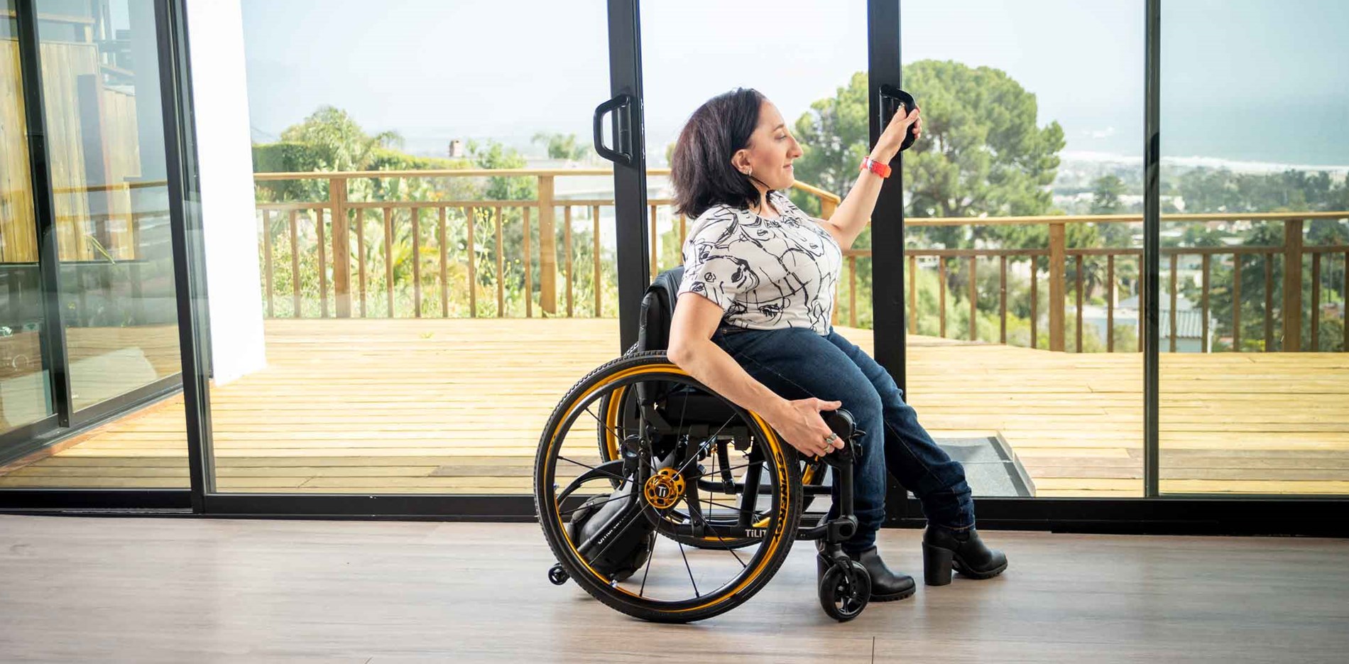 A woman using a manual wheelchair with SmartDrive slides open the glass door leading onto a large wooden patio overlooking a green cityscape.