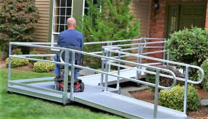 A man, viewed from the back, drives his scooter up a metal modular ramp towards the entrance to his house.