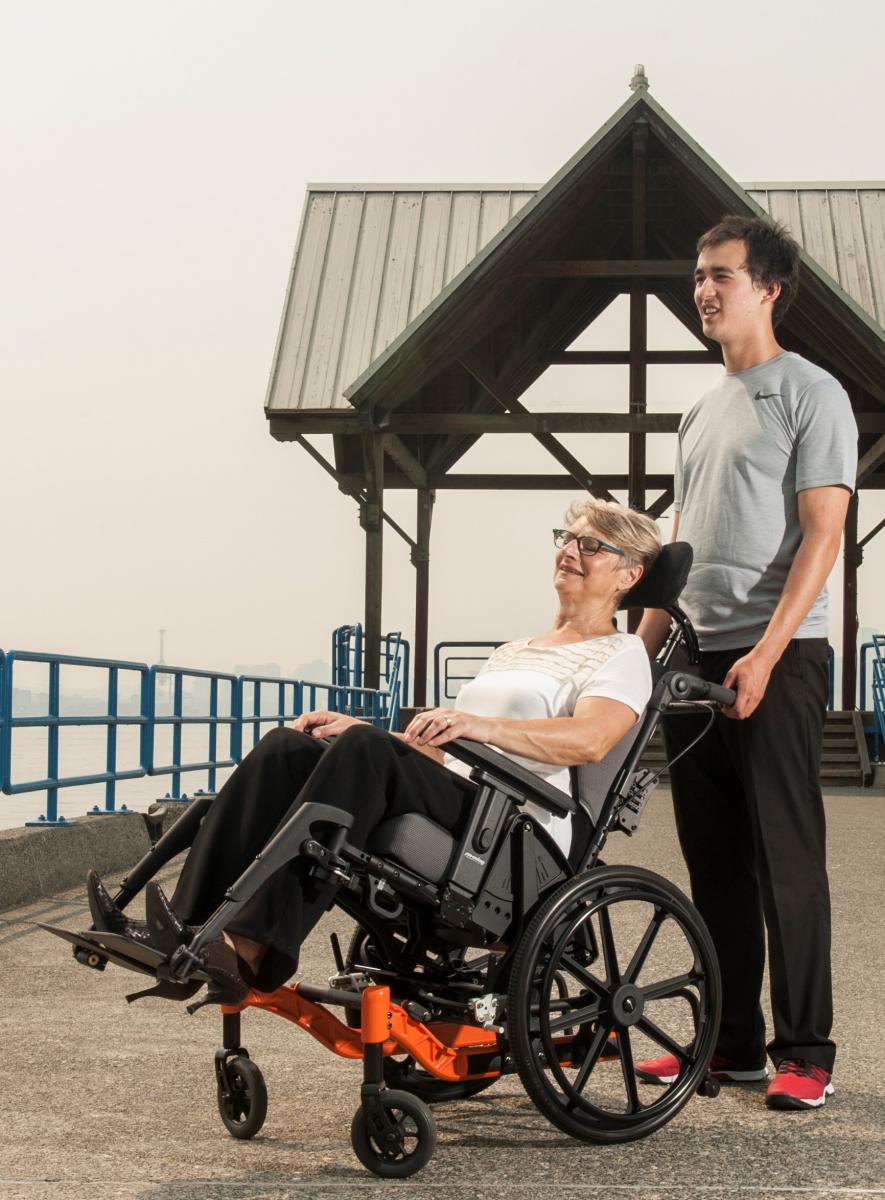 A woman sits tilted in a wheelchair on a pier. A smiling man stands behind her.